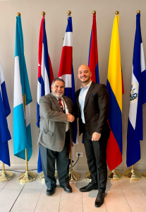 Dos representantes institucionales se saludan frente a banderas de países centroamericanos y del Caribe