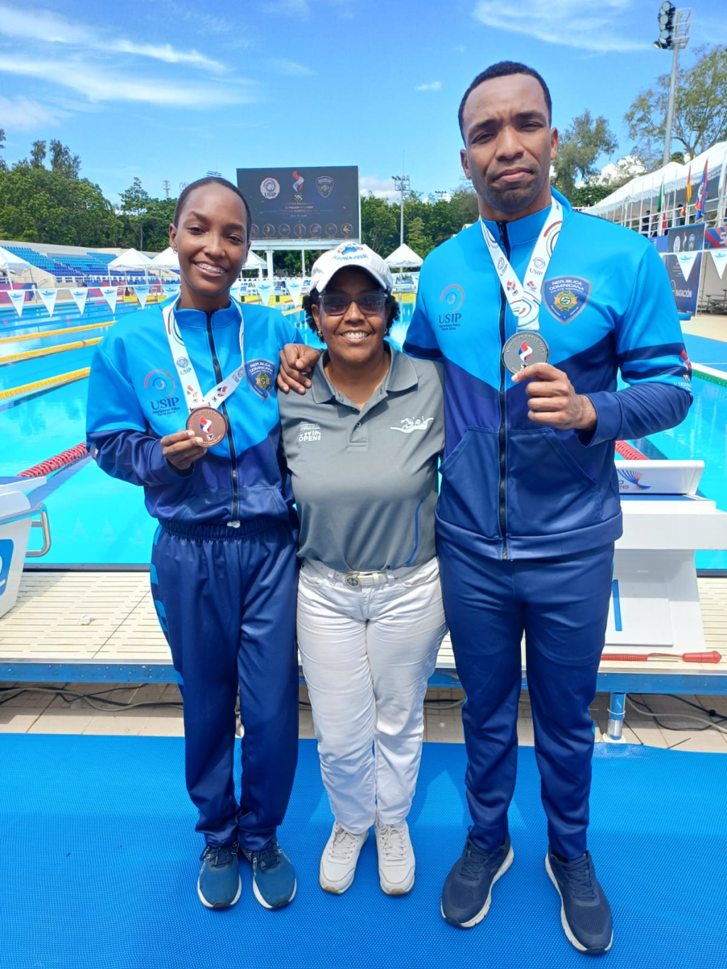 Madre junto a dos atletas dominicanos de natación que exhiben medallas en piscina de competencia